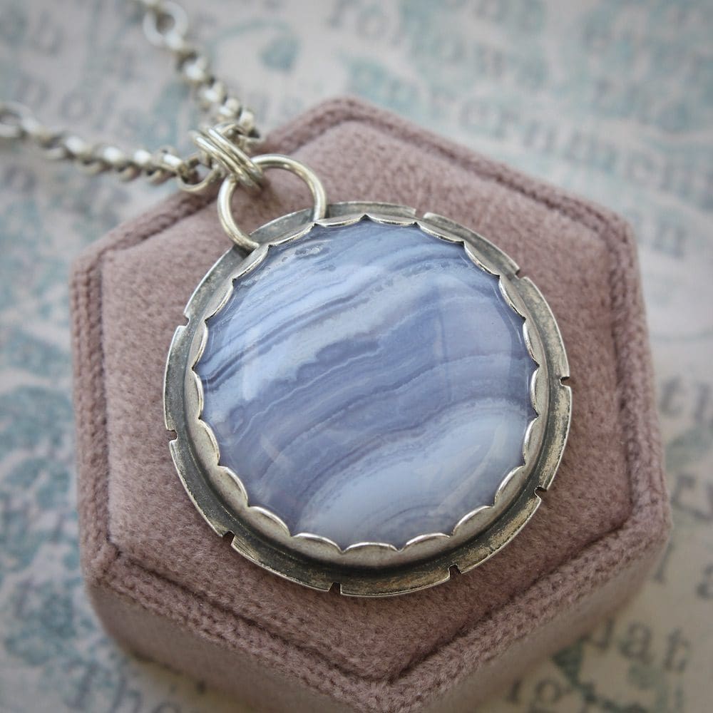 A circular blue lace agate and sterling silver gemstone pendant on a silver chain, on a hexagonal pink velvet box lid and a cream and blue background with old text on it.