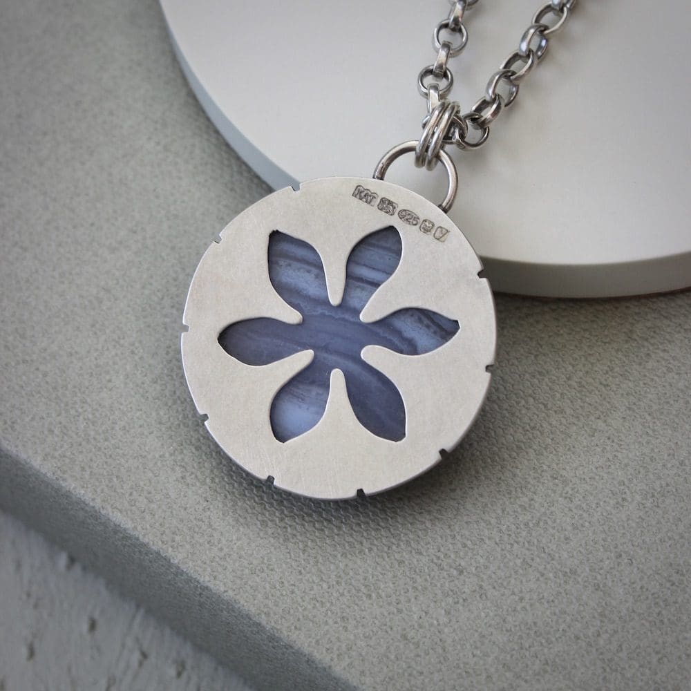 A circular blue lace agate and sterling silver gemstone pendant on a silver chain, propped against a blue ceramic coaster showing a cut-out flower on the reverse of the design and a hallmark.
