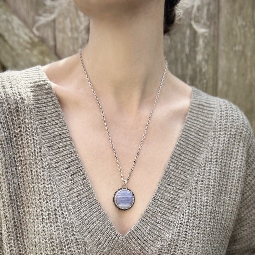 A circular blue lace agate and sterling silver gemstone pendant worn on a silver chain by a woman wearing a sage green knitted jumper and standing in front of a wooden gate.
