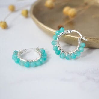 pair of blue-green beaded hoope earrings rest on small brass dish with dried flower stems all on white background