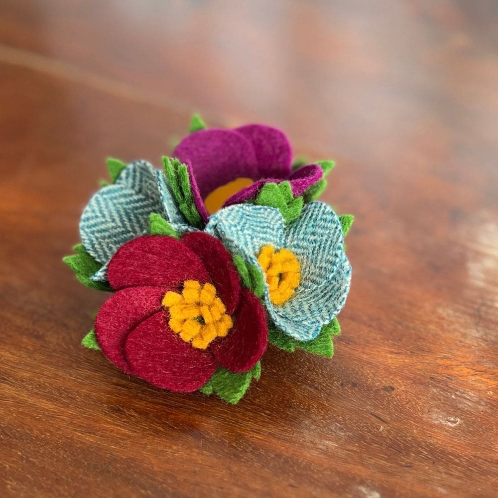 A 1940s style, handmade brooch, sits on a wooden background. The brooch is made up of four small, wool felt flowers that have been hand stitched together to form a posy.