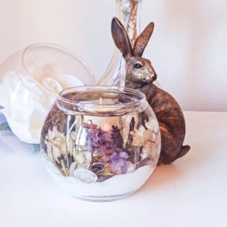 Luxury lavender scented candle. Fishbowl soy and gel wax tumbler with shells and dried flowers in shades of purple and white is on a white dresser with a large white flower, glass vase and a bronze hare ornament.