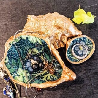 Oyster shell decoupaged with William Morris Seaweed design and gold gilded edges is next to a small clam shell with the same design. The shells are on a dark back ground and are draped in jewllery.