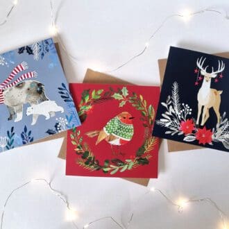 Three illustrated Christmas cards are displayed on a white background with white fairy lights around them. The card on the left features two polar bears, the larger of which wearing a red and white stripy scarf, on a purple background with foliage at the edges. the middle card features a robin in a green jumper, in the middle of a wreath, on a red background. the final card features a stag with stars hanging from it's antlers with foliage below, on a deep blue background.
