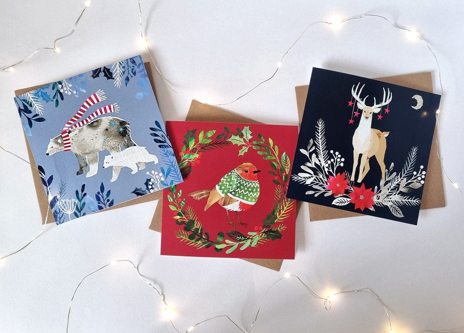 Three illustrated Christmas cards are displayed on a white background with white fairy lights around them. The card on the left features two polar bears, the larger of which wearing a red and white stripy scarf, on a purple background with foliage at the edges. the middle card features a robin in a green jumper, in the middle of a wreath, on a red background. the final card features a stag with stars hanging from it's antlers with foliage below, on a deep blue background.