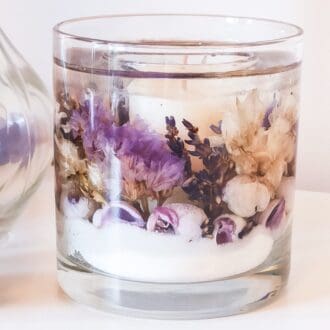 Soy and gel wax scented candle on a pale surface next to a glass vase. The scented tumbler is decorated with white sand, shells and dried flowers, including lavender, in shades of purple and white.