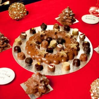 chocolate wreath set on a table surrounded and filled with chocolate truffles, with four christmas trees set around it.