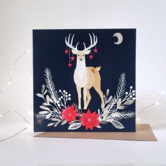 An illustrated Christmas card displayed on a white background with white fairy lights around it. The card features a stag with stars hanging from it's antlers with foliage below, on a deep blue background.
