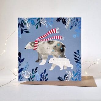 An illustrated Christmas card displayed on a white background with white fairy lights around it. The card features two polar bears, the larger of which wearing a red and white stripy scarf, on a purple background with foliage at the edges.