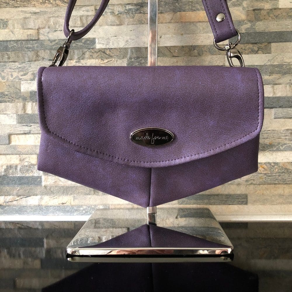 Discover a handcrafted clutch or shoulder bag made from vegan leather in a rich grape purple, featuring an oval, handcrafted metal label. Its unique design boasts a distinctive v-shaped base.