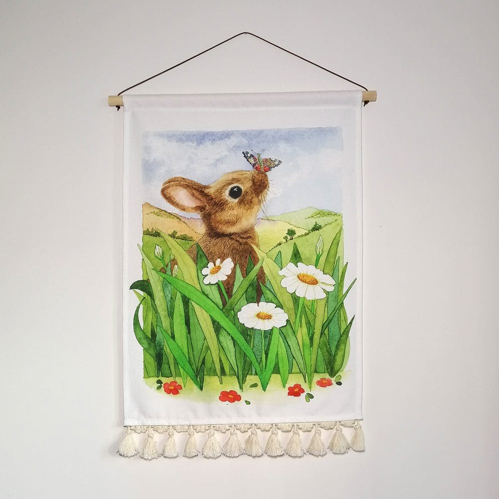 Young will rabbit sat amongst the long grasses and daisies with a butterfly friend on her nose. Printed on heavyweight cotton, the wall hanging has a wooden pole hanger, fine brown waxed cotton chord, and a decorative pale cream cotton tassel border giving a contemporary look to the lovely wall decor.