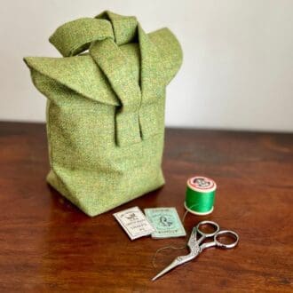 a small two handled bag made from green wool tweed sits on a wooden top next to some vintage, scissors, thread and an old packet of needles.