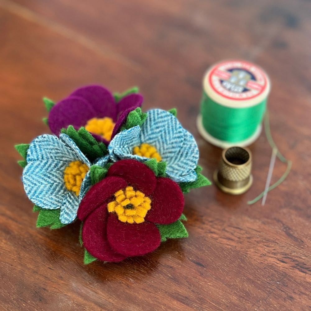 A small posy of wool felt flowers, sits on a wooden background next to a vintage cotton reel and thimble.