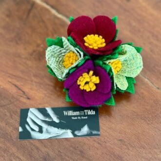 Photo shows a brooch of two green, one purple and one red wool felt flowers. The brooch sits on a wooden top next to a card saying the item was handmade by the brand William and Tilda.