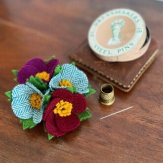 A brooch of four small, wool felt flowers sits next to some vintage haberdashery on a wooden table top.