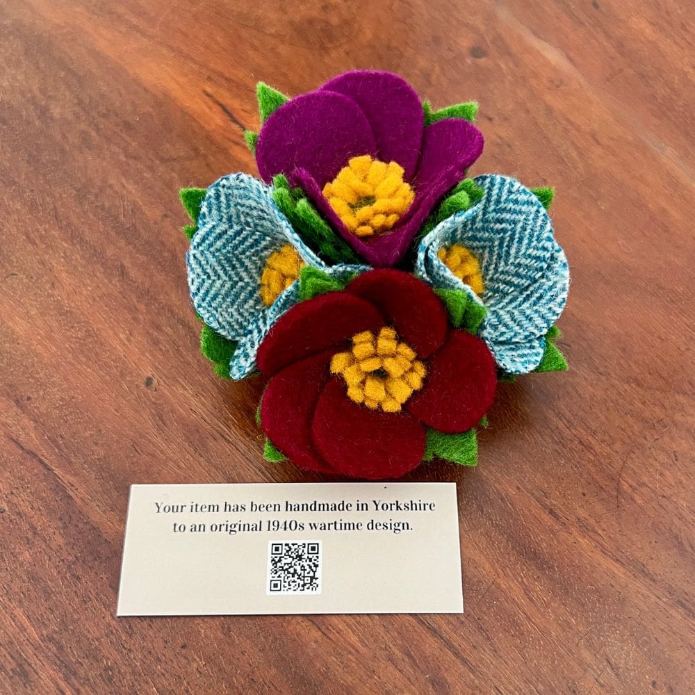 Photo shows a four flowers felt brooch on a wooden top. A card sits in front of the brooch saying 'handmade in yorkshire from an original wartime design'.