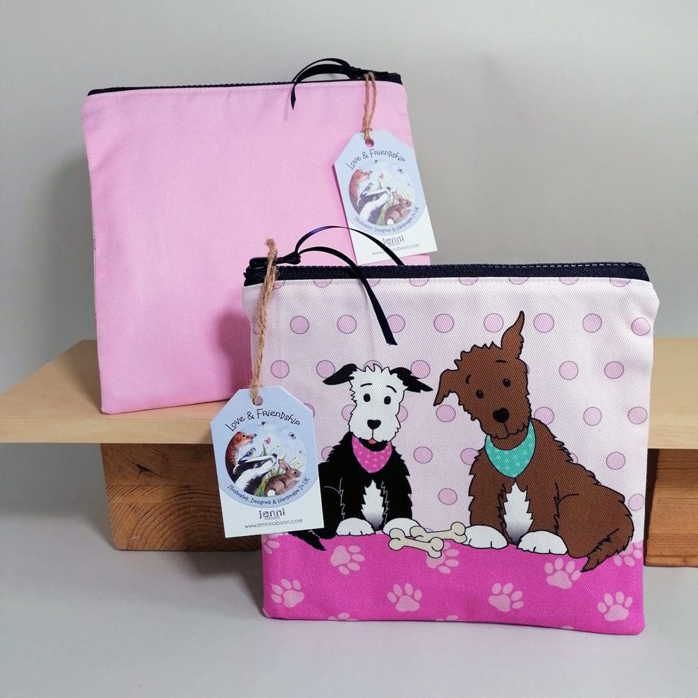 Flat, cotton toiletries bag featuring two dogs in bandanas with their bones. Various shades of pink, spotty background, paw print base and a pale pink back.