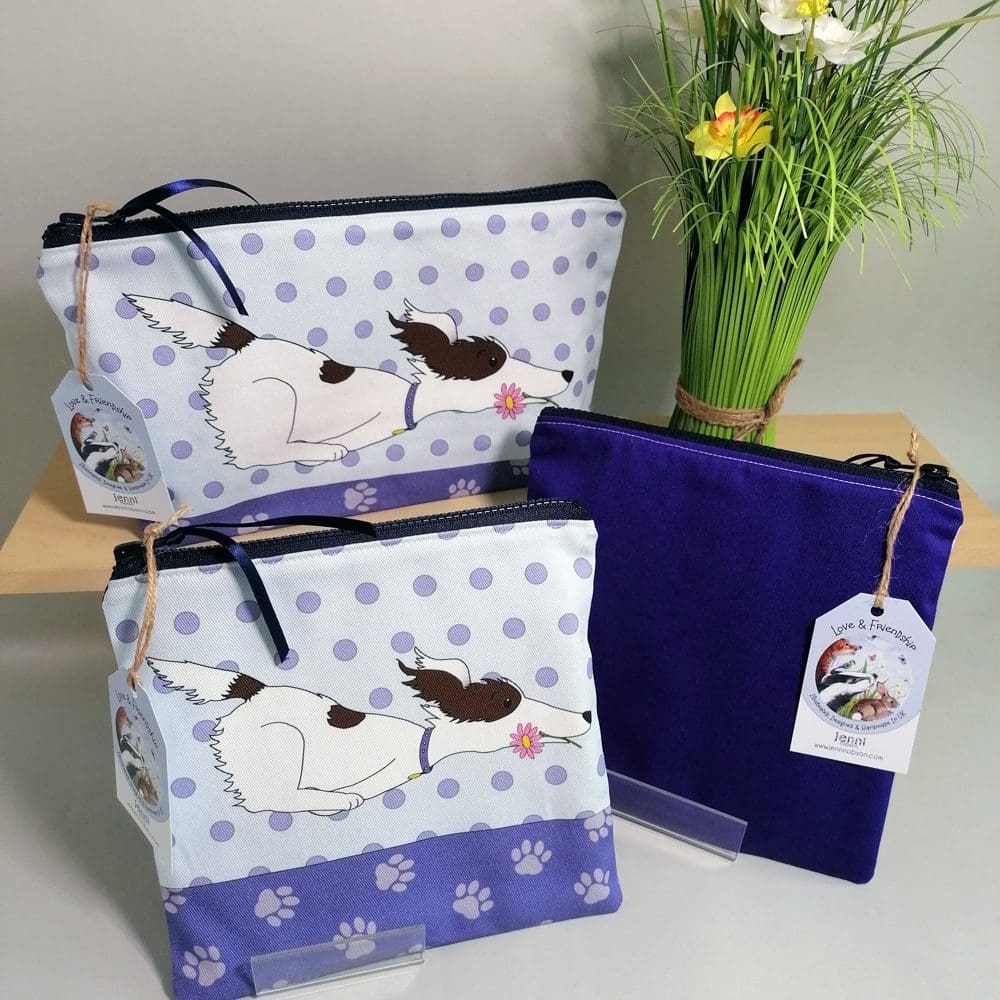 Large and medium cotton toiletries bag featuring a zooming dog with a pink flower in its mouth. Various shades of purple and pale blue, spotty background, paw print base and a dark purple back.