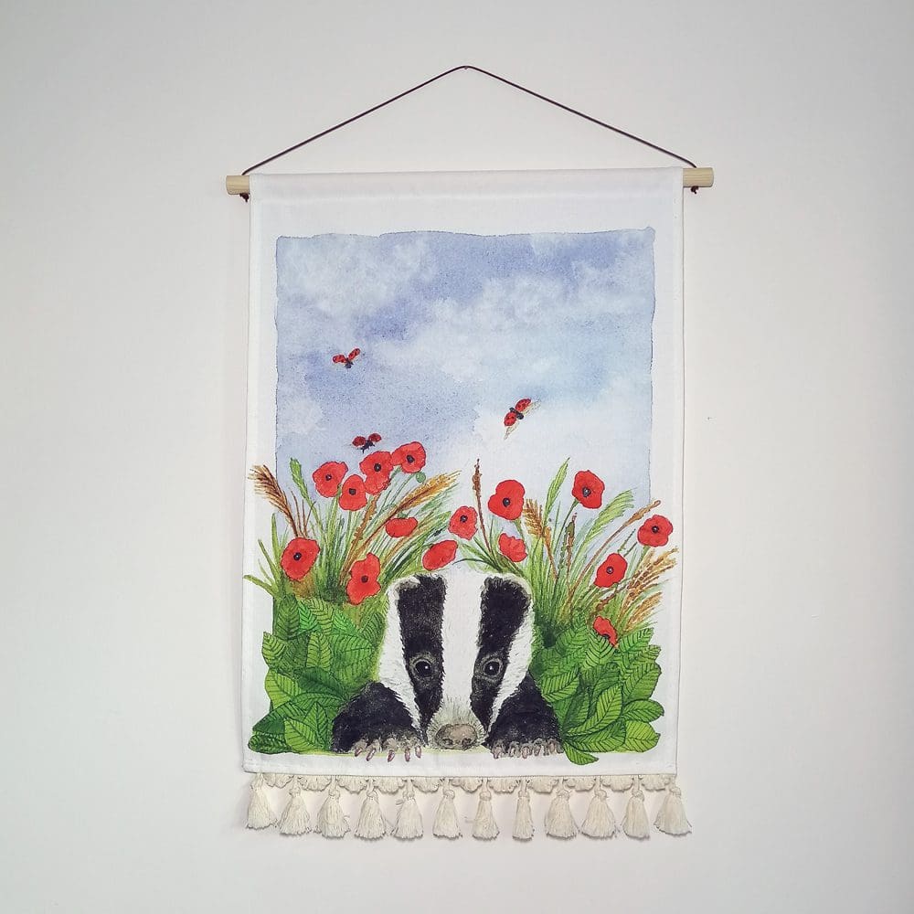 Contemporary cotton wall hanging with decorative pale cream tassel details, a wooden hanging pole and waxed brown cotton hanging chord. Featuring a young badger peeking out of the poppies and leaves