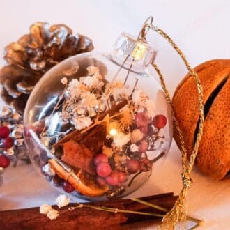 Silver and pink berry botanical glass christmas bauble is placed next to cinnamon sticks, a dried orange and fir cone.