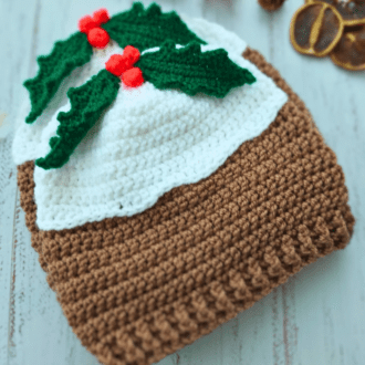Novelty crochet Christmas pudding beanie hat garnished with holly leaves and red berries. The top section is white to mimick the rum or brandy sauce, followed by a mocha brown for the pudding section.