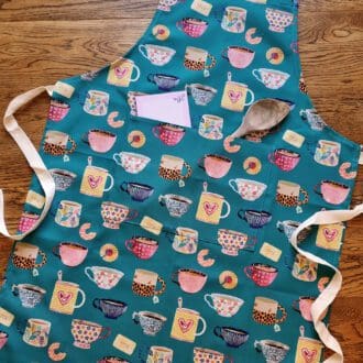 Teal coloured cotton apron with adjustable neck fastening and pocket. Printed with illustrations of colourful tea cups and biscuits.