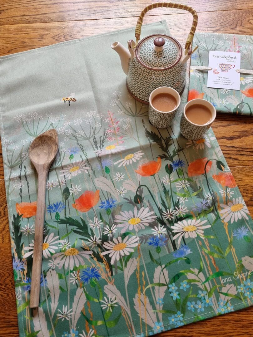 A cotton tea towel printed with a wildflower illustration. The tea towel is laid out on the table with teapot and two small cups of tea to the right hand side.