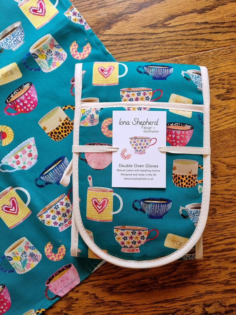 Teal coloured oven gloves with towelling reverse, printed with illustrations of colourful tea cups and biscuits.