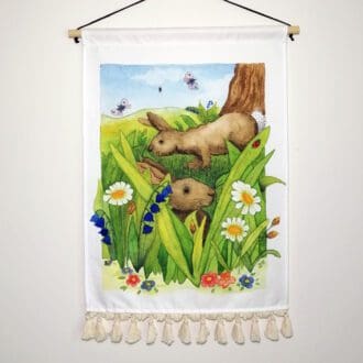 Two brown rabbits amongst the long grass and wild flowers. Contemporary cotton wall hanging with decorative tassel details, a wooden hanging pole and waked cotton hanging chord.