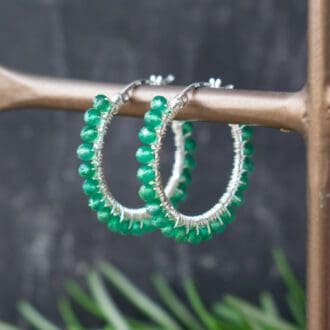 green beaded hoop earrings hung on brass stand with foliage in background