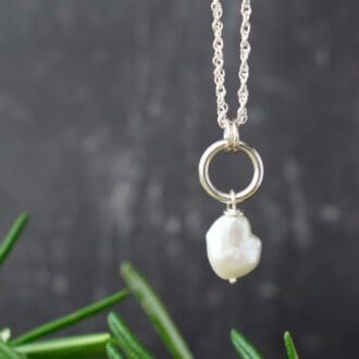 Silver necklace featuring an irregular keishi pearl hung from a small silver hoop. Dark background with greenery.