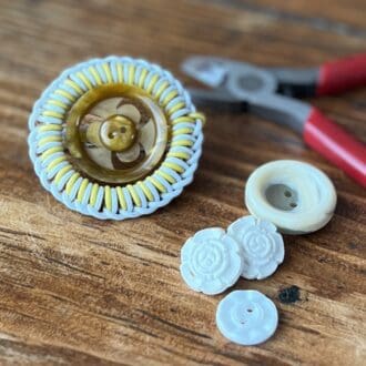 A circular vintage style brooch with an amber coloured vintage button centre sits on a wooden top surrounded by pliers and an assortment of white buttons.