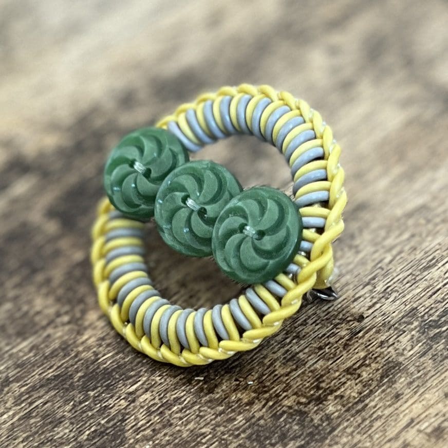 Photo shows a circular yellow and grey brooch with three small green buttons forming the centre. The brooch sits on a distressed wooden top.