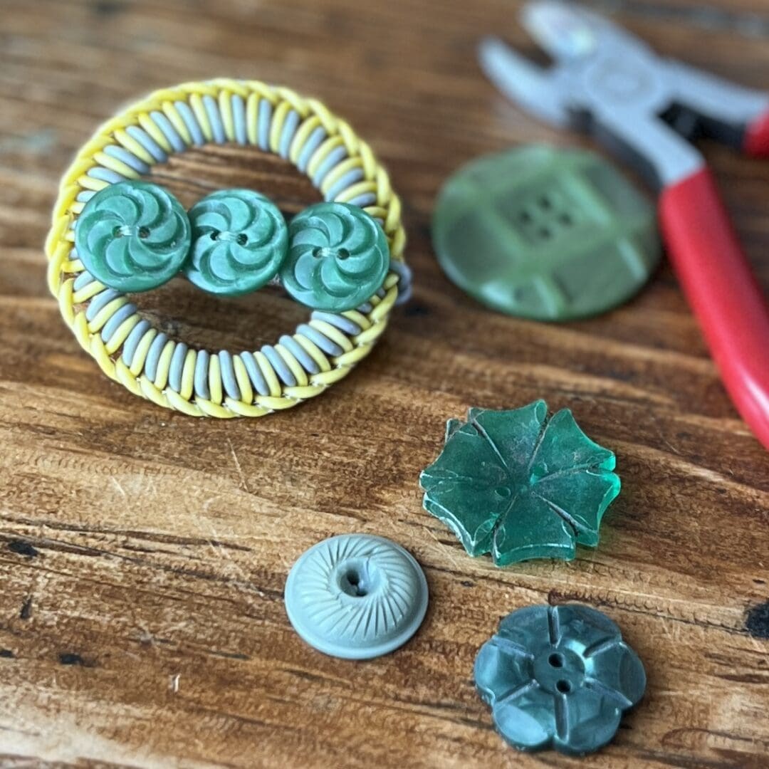 Circular grey and yellow coloured wire brooch with three green textured buttons sitting across the middle in a line. The brooch rests on a wooden table surrounded by pliers and other green buttons.