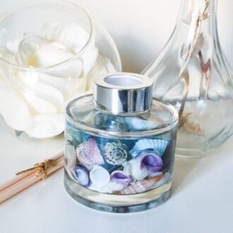 Circular blue sea ocean diffuser 165ml, contains real shells and blue oil, on white background surrounded by decorative glass and flowers.