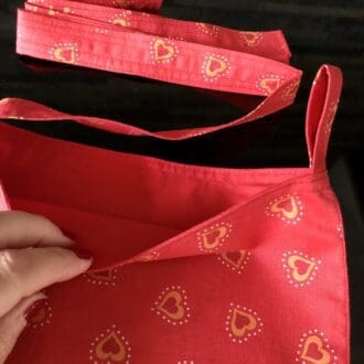 Adjustable cross body style mastectomy drain bag with a red fabric covered with hearts, this has a red cotton lining, on a reflective black surface