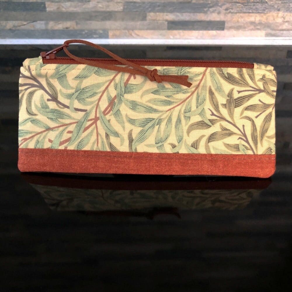 A zipped pencil case a green and brown Willow design by William Morris with faux suede pull, pale pink lining on shiny black surface.
