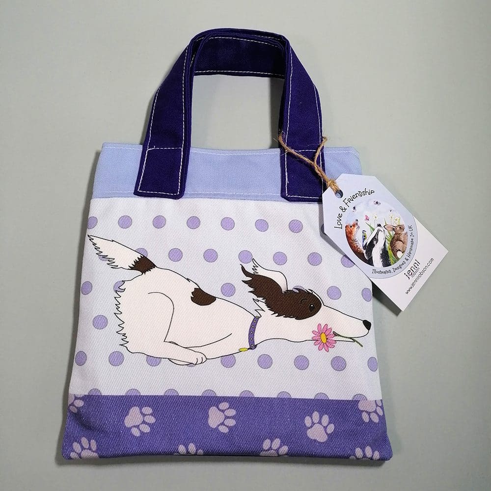 Front view of the mini bookbag in dark purple and pale blue cotton fabric. Featuring a running white dog with white spots carrying a flower in his mouth. Lined with water resistant lining