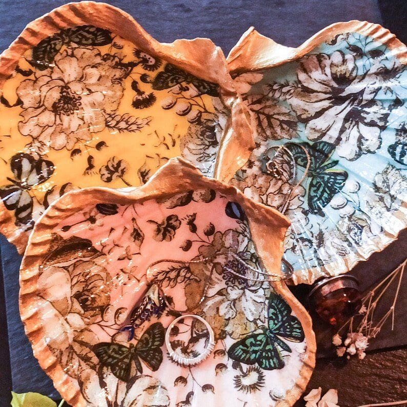 Set of three vintage design decoupage scallop shell trinket dishes in pink, yellow and blue. The dishes are on a dark slate background with jewellery strewn around them.