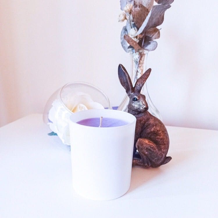 Lavender scented soy wax candle. The lavender coloured candle comes in a white cylindrical candle jar and is placed on a white vanity unit surrounded by dried flowers and a hare ornament.
