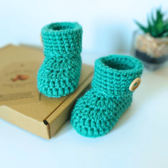 A pair of sage green crochet baby booties with a folded cuff that is finished off with a wooden 'handmade with love' button. Sizes available are newborn 0-3 and 3-6 months with a variety of colours to choose from