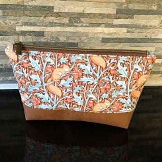 An extra large cosmetic bag in Tilda Sleepy Squirrels brown zip and faux suede pull. A base of chestnut brown faux leather. Standing on a reflective black surface.