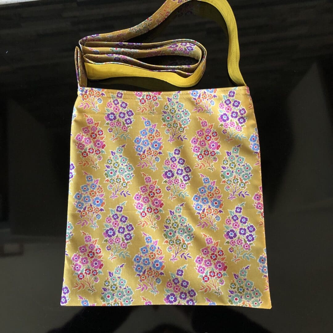 Adjustable cross body style, mastectomy drain bag in mustard yellow fabric with stylised flowers mustard cotton lining, lying on a reflective black surface.