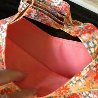 Adjustable cross body style, mastectomy drain bag in orange fabric with bunches of colourful summer flowers and a pale orange cotton lining, lying on a reflective black surface.