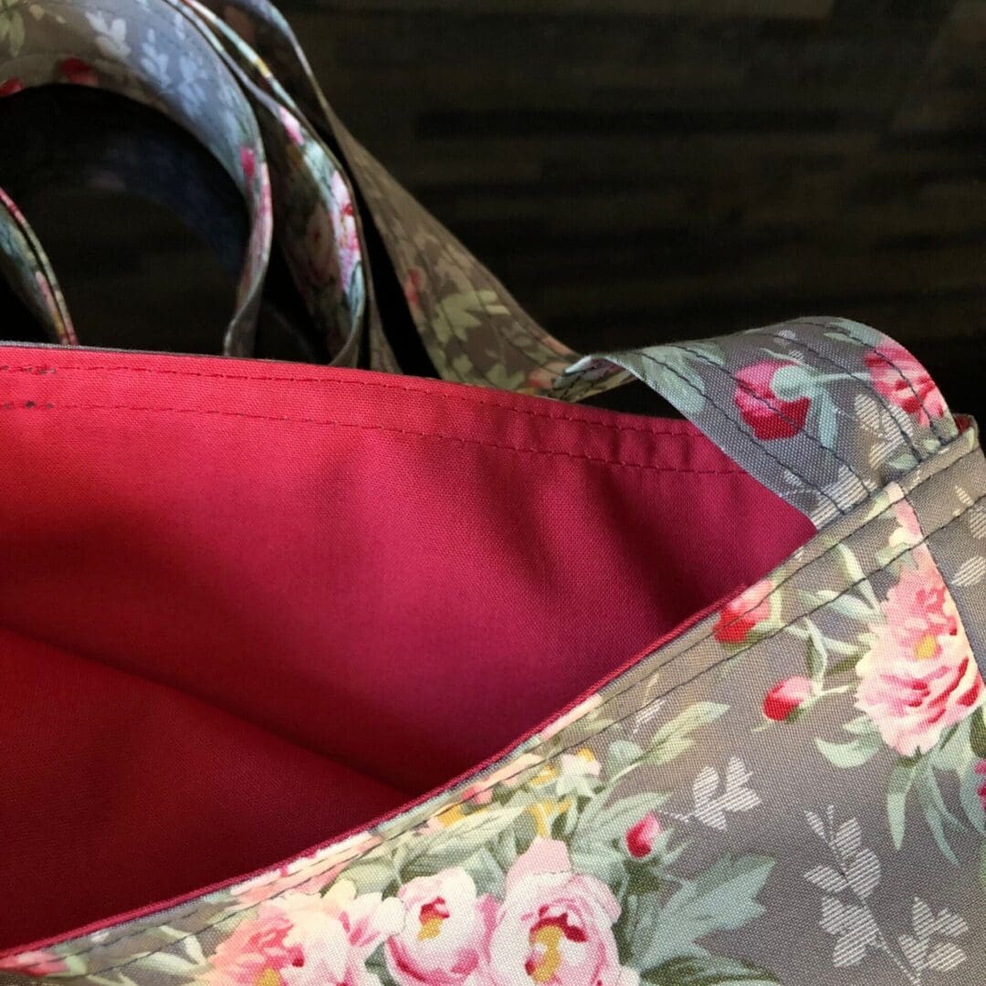 Adjustable cross body style, mastectomy drain bag in grey Tilda fabric with clusters of pink roses and a dusky pink cotton lining, lying on a reflective black surface.