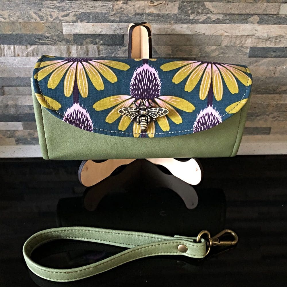 Ladies clutch wallet in olive faux leather featuring an echinacea print flap with large brass bumblebee clasp on a stand
