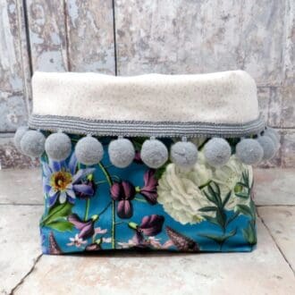 Fabric storage bin made in a turquoise floral velvet and with a grey pom pom trim.