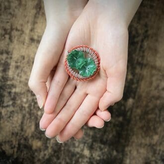 Photo shows two hands crossed with a 1940s style creative wire worked brooch in one palm. The brooch is made from orange and grey wire twisted in an intricate pattern with two green, vintage buttons forming the centre of the circular brooch.