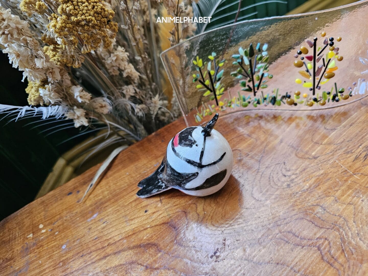 Ceramic great spotted woodpecker on a wooden table with dried flowers and glass art in the background