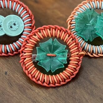 Three circular brooches made from twisted coloured wire sit on a wooden tabletop. Each of the brooches has a vintage green button set in the centre.